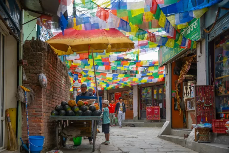 Kathmandu alleyway draped in prayer flags, with a vendor and child by a fruit cart.