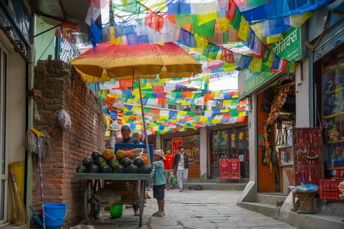 Kathmandu alleyway draped in prayer flags, with a vendor and child by a fruit cart.