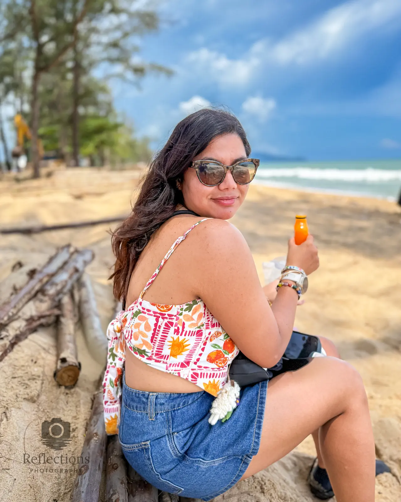 Woman sitting on Sai Kaew Beach with orange drink, Andaman waves and casuarina trees in the background.